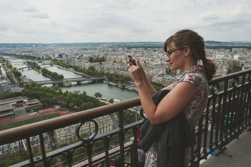 Woman photographing on mobile phone while standing by railing against sky