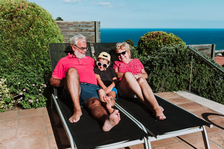 Smiling grandmother and granddad lying on deck chairs together with adorable boy and relaxing during summer weekend