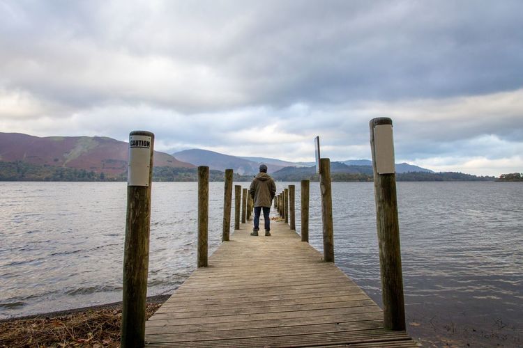Rear view of mature man standing on pier over lake against cloudy sky