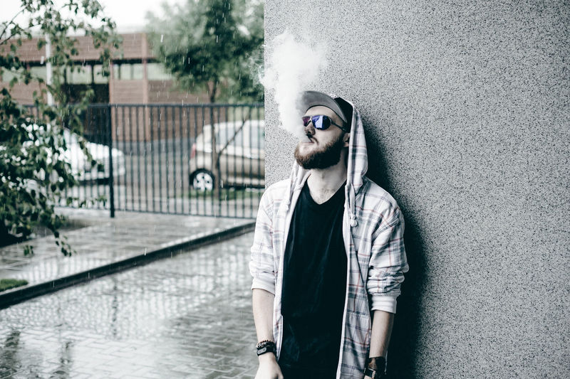 Close-up of young man smoking while standing by wall during rain