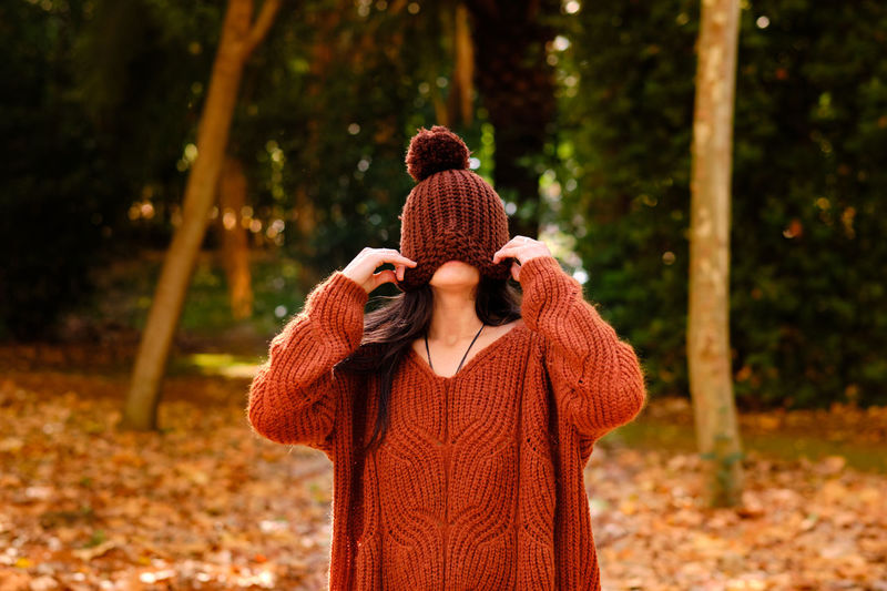 Unrecognizable female in knitted hat covering face wearing a warm sweater while standing in forest in cold autumn day