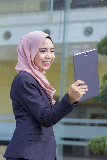 Happy businesswoman using digital tablet while standing against building