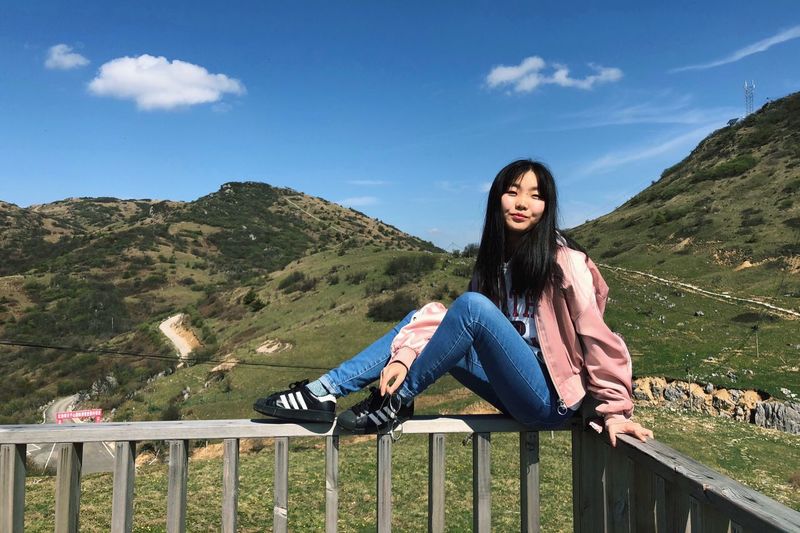 Portrait of smiling young woman sitting on railing against mountains