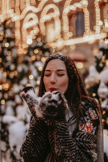 Young brunette woman in winter warm clothes in christmas street scenery blowing snow from her hands. 