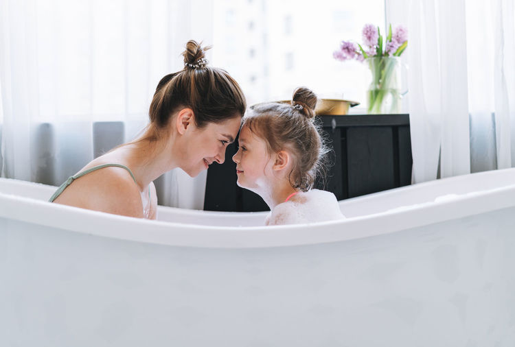 Young mother with little tween girl daughter in pajamas having fun in the bath with foam at home