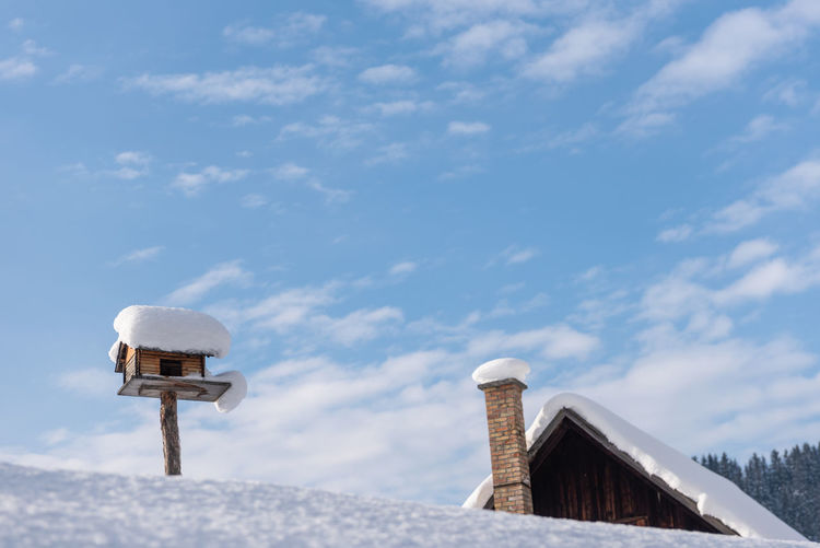 Homemade wooden bird's feeder in winter, under snow. blue cloudy sky, house at the background.