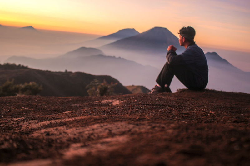 Man sitting on mountain against sky during sunset