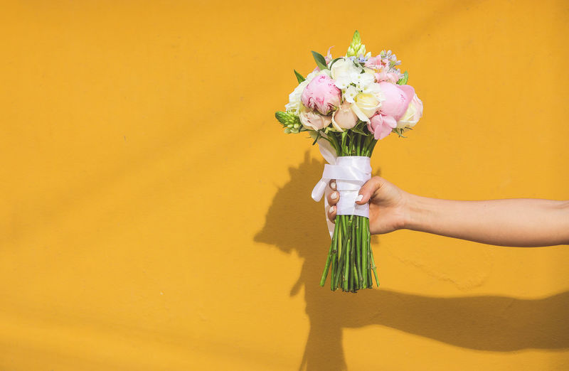 Cropped hand of woman holding bouquet against yellow wall