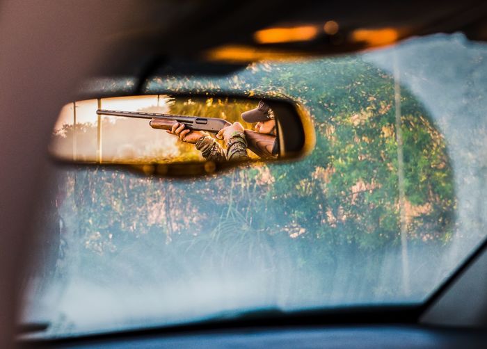 Hunter aiming with shotgun reflecting in rear-view mirror of car