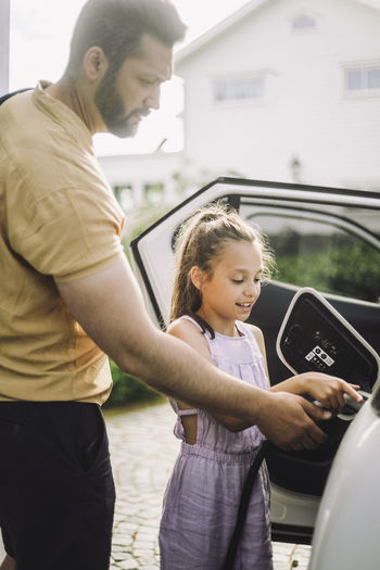 Girl learning to charge electric car while standing by father