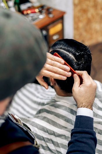 High angle of male barber with comb brushing hair of unrecognizable client while making hairstyle in modern barbershop