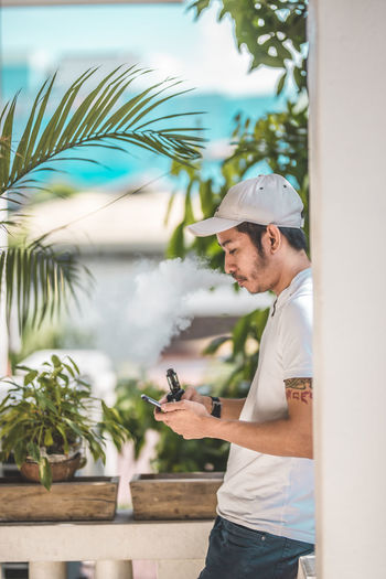 Side view of man smoking electronic cigarette while using smart phone on balcony