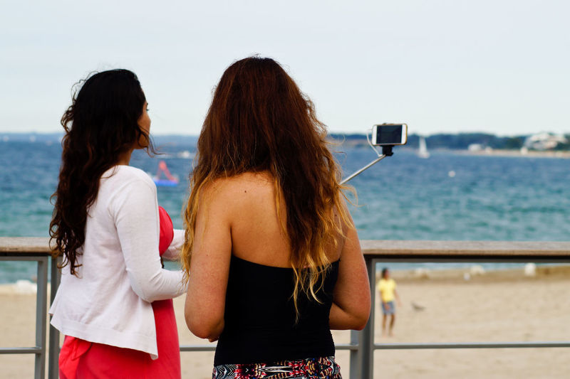 Female friends taking selfie while standing by railing at beach against clear sky