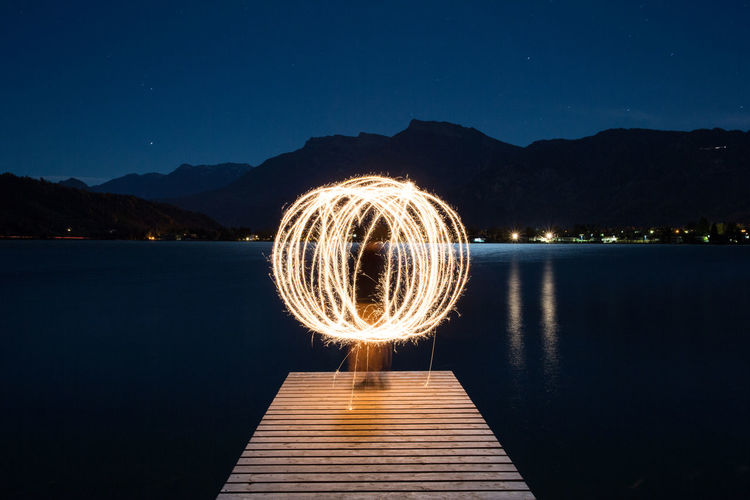 Person spinning illuminated wire wool while standing on pier against lake at night