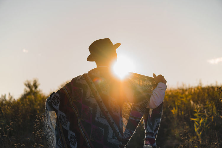 Man in poncho and hat walks away against the setting sun, rural area