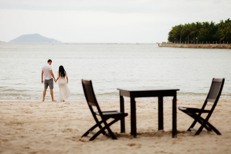 Rear view of couple at beach with table and chairs in foreground