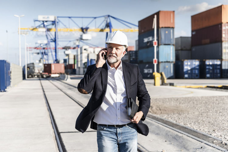 Businessman at cargo harbour, wearing safety helmet, using smartphone