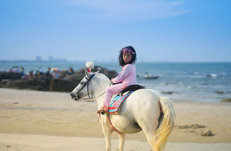 Full length of girl sitting on horse at beach against sky