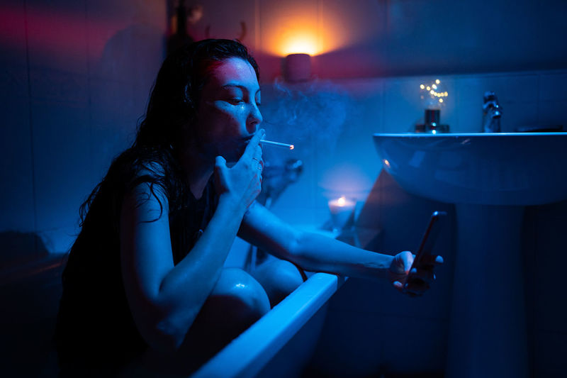 Young nicotine addicted woman with smartphone smoking cigarette, sitting in bathtub under neon light
