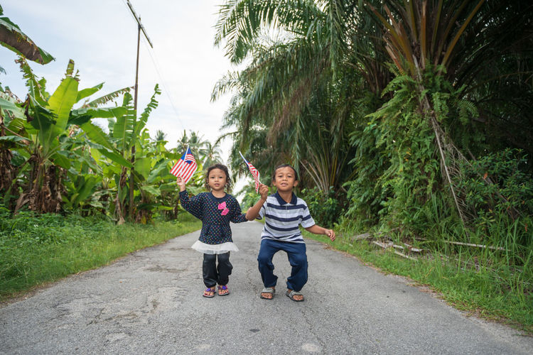 Siblings holding malaysian flag while standing on road amidst plants