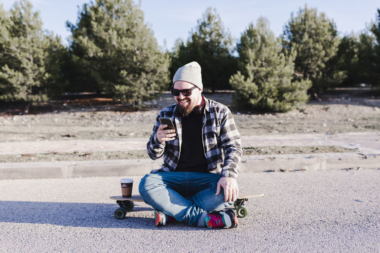 Modern young man sitting on a longboard looking at his mobile phone
