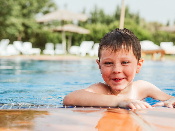 Delighted cute child with wet hair leaning on poolside and looking at camera while having fun during summer weekend