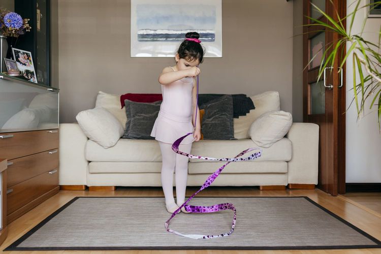 Focused cute little brunette girl in leotard and tights while spinning ribbon during rhythmic gymnastic practice training in cozy living room at home
