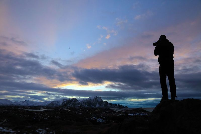 Rear view silhouette of man photographing while standing on rocks against sky during sunset