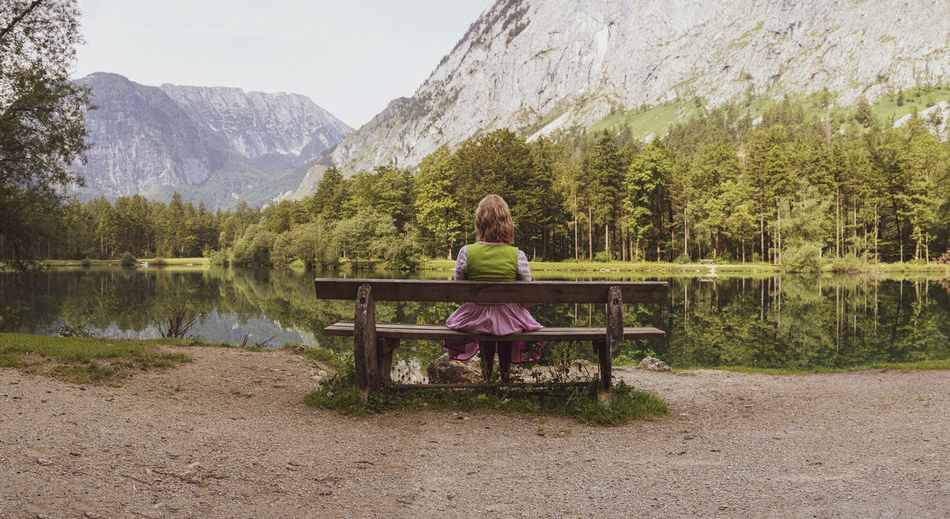 Rear view of woman sitting on bench by lake against mountains