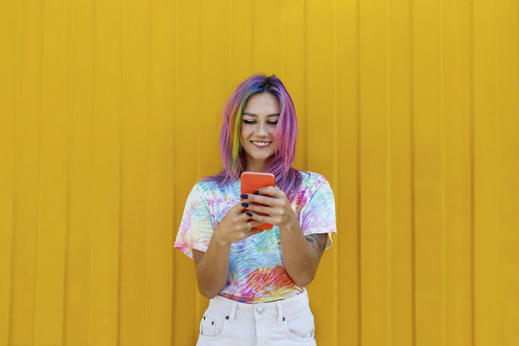 Smiling young woman wearing tie dye t-shirt using smart phone in front of yellow wall