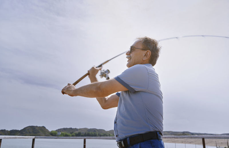 Senior man fishing with fishing rod by sea on sunny day