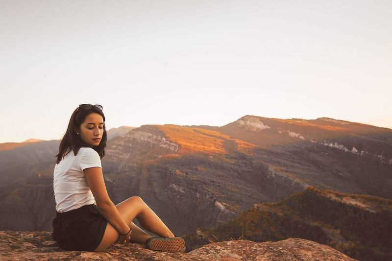 Portrait of young woman sitting on mountain against clear sky