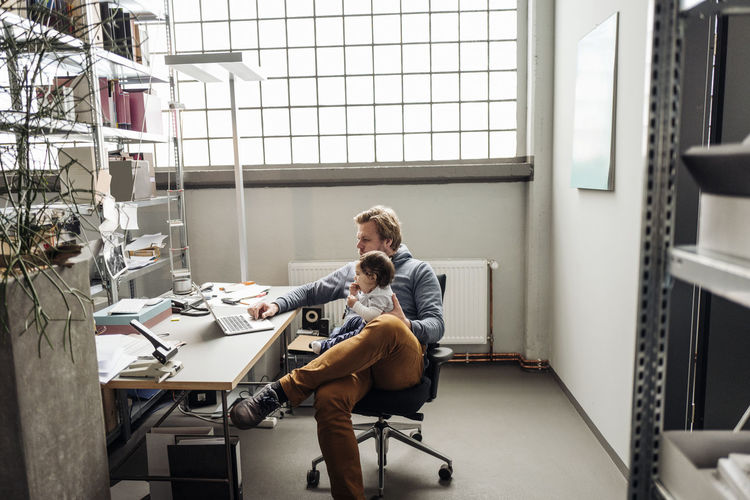 Man with baby on lap using laptop at desk in a factory
