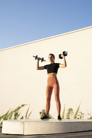 Sportswoman flexing muscle with dumbbell standing on pedestal against wall during sunrise