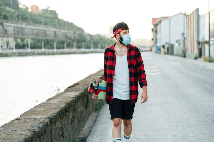 Young male skater in surgical mask and with longboard walking along promenade during coronavirus pandemic and looking away