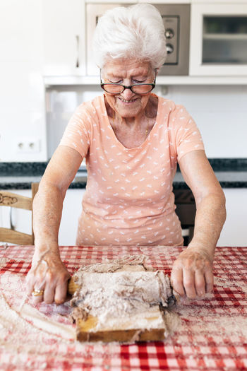 Cheerful old female with gray hair in eyeglasses rolling out dough on cutting board at table in house