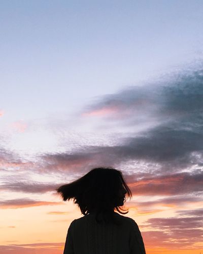 Rear view silhouette of woman tossing hair while standing against sky during sunset