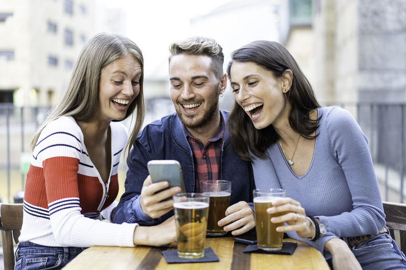 Portrait of smiling friends using mobile phone while sitting on table
