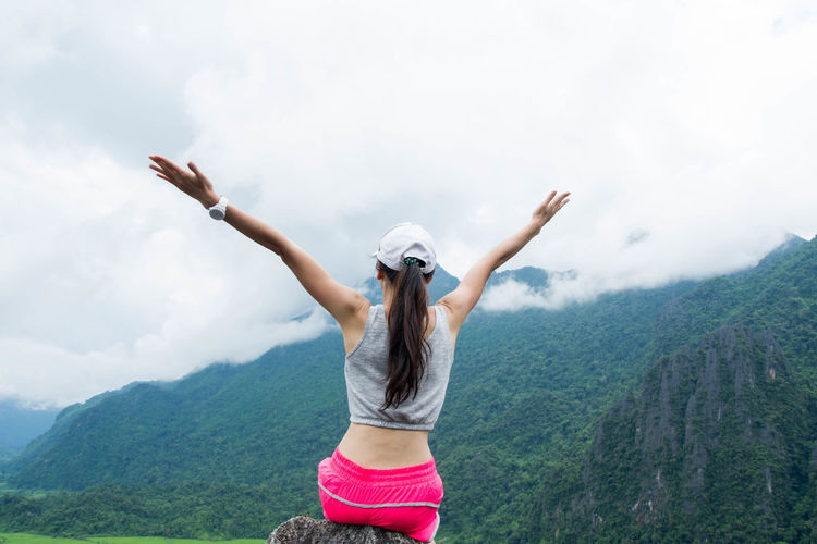 Rear view of woman with arms raised sitting on mountain against sky