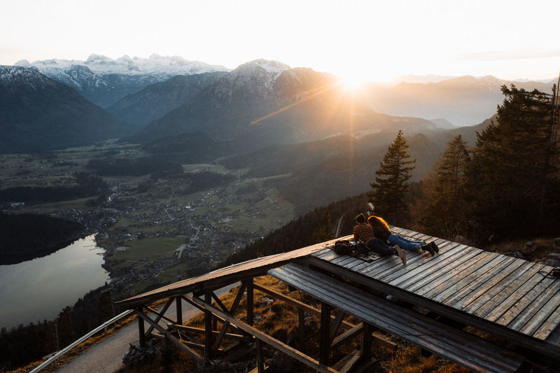 Back view of unrecognizable couple of travelers lying on wooden terrace and enjoying sunrise in highlands in foggy morning