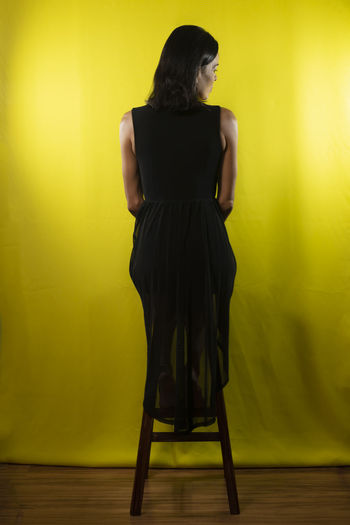 Portrait of model sitting on wooden bench with back in black dress against yellow background 