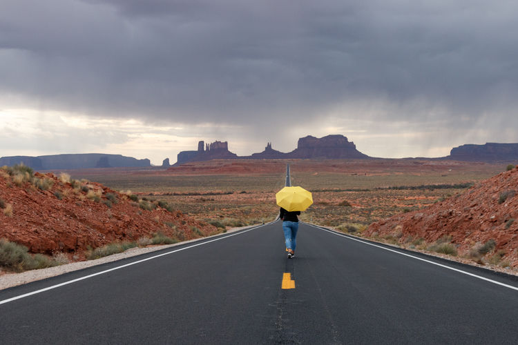 Woman with umbrella walking on road leading towards monument valley
