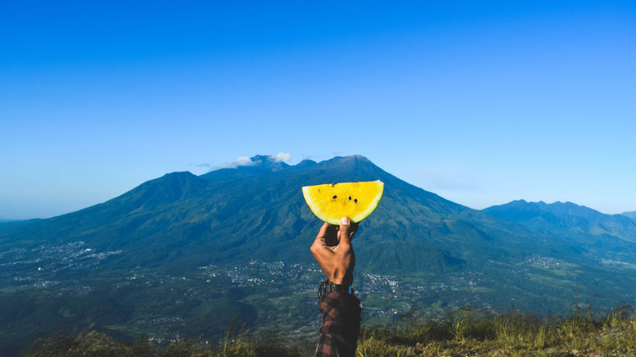 Cropped hand of man holding watermelon slice against blue sky