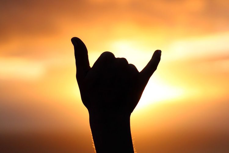 Close-up of silhouette hand gesturing shaka sign against orange sky