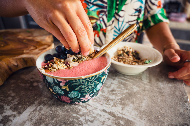 Crop anonymous female putting crunchy sweet granola into tasty pink smoothie bowl with blueberries during breakfast in kitchen at home