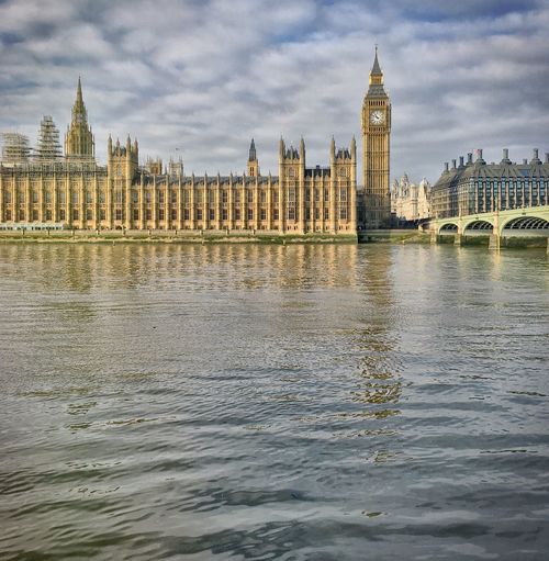 Houses of parliament by thames river against cloudy sky