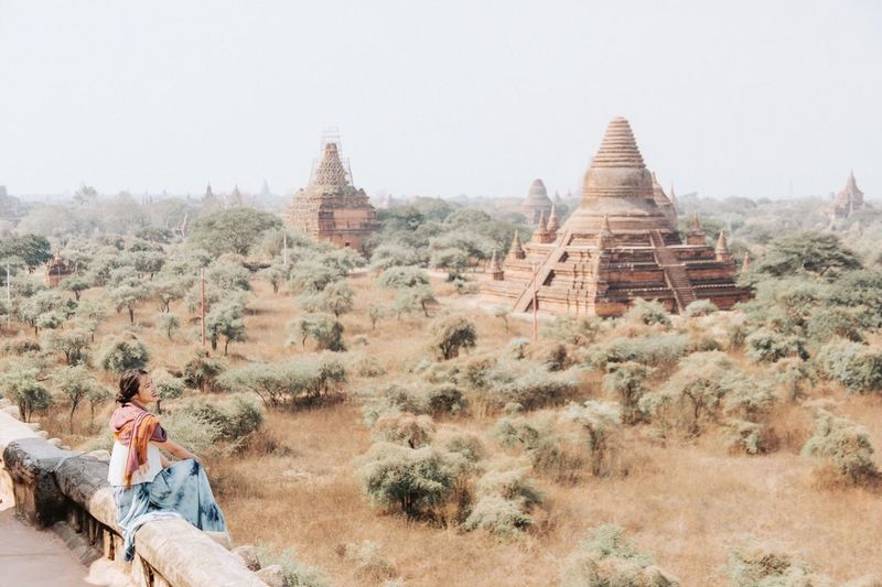 Woman sitting on retaining wall by temple against clear sky
