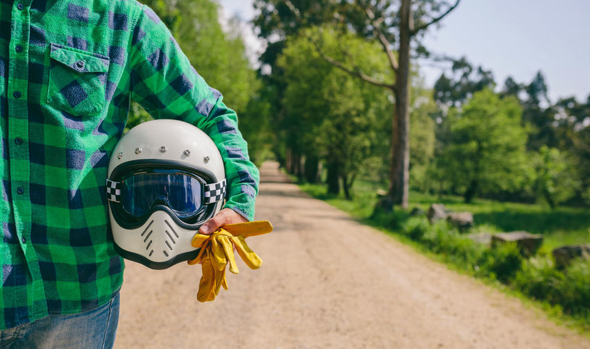 Midsection of man holding crash helmet and gloves while standing on dirt road against trees in forest