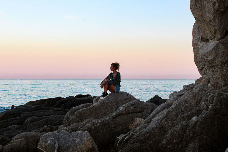 Woman sitting on rock by sea against sky during sunset