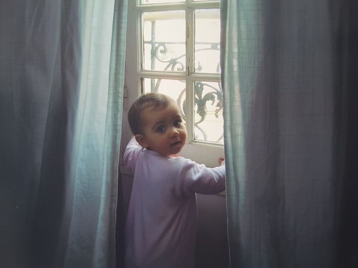 Portrait of cute baby girl standing by window at home